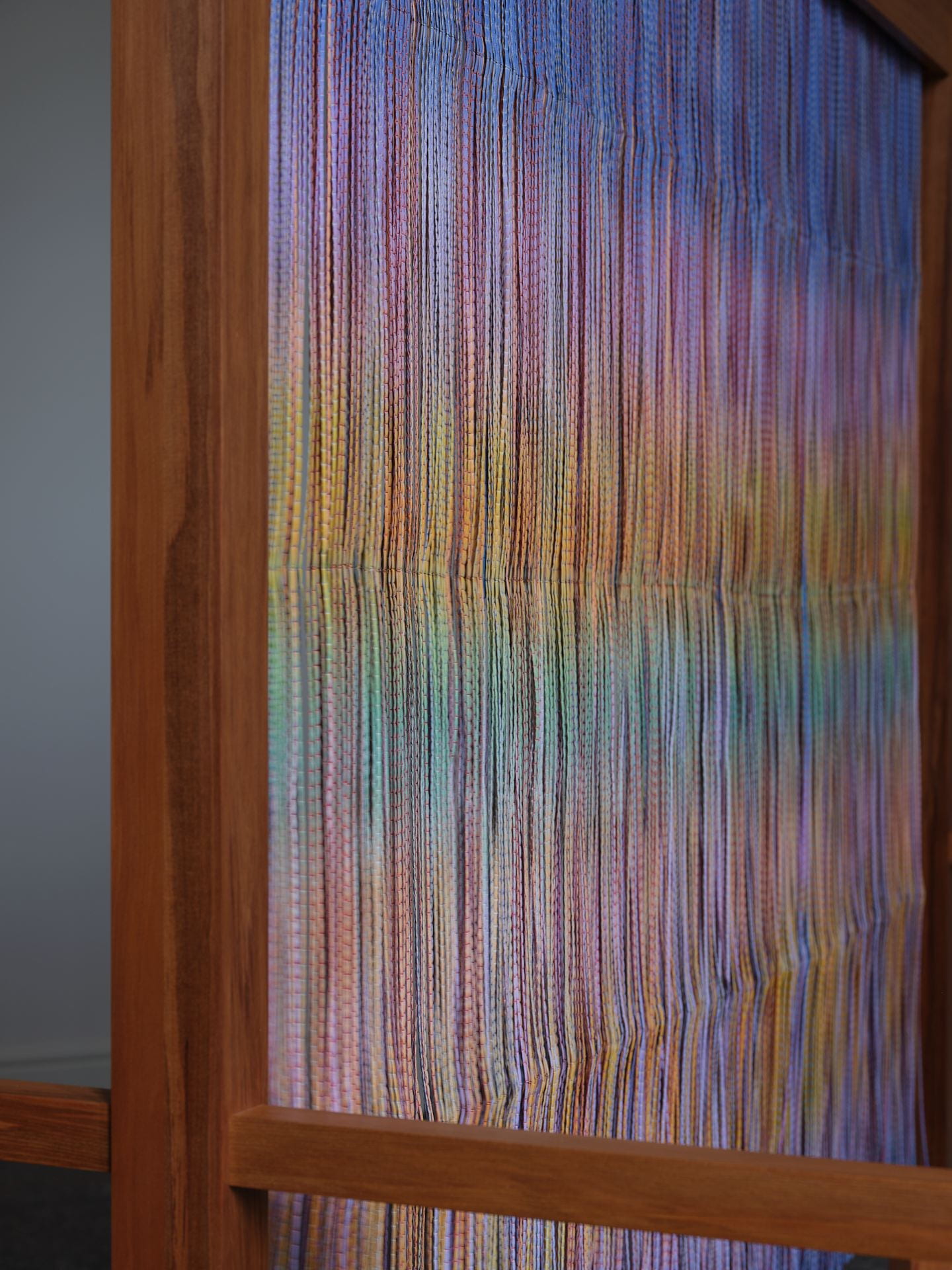 Close-up of a wooden structure with a veil hanging from it. The veil is made up of multicoloured strands in purples, oranges, greens and pinks.