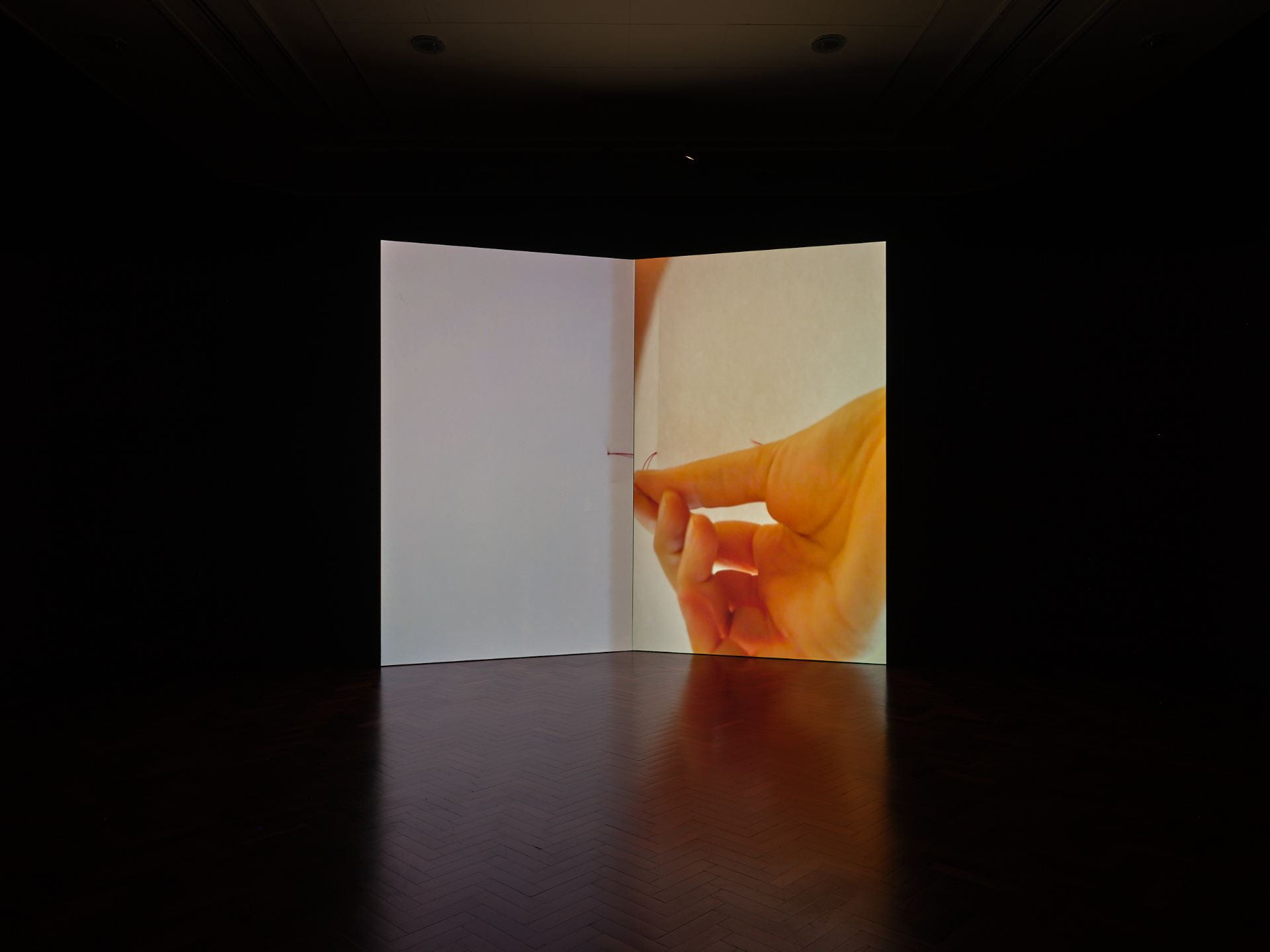 A film work is projected onto a structure shaped like an open book. The film shows a person's hand sewing a book-binding seam in red thread across the centre of the structure.
