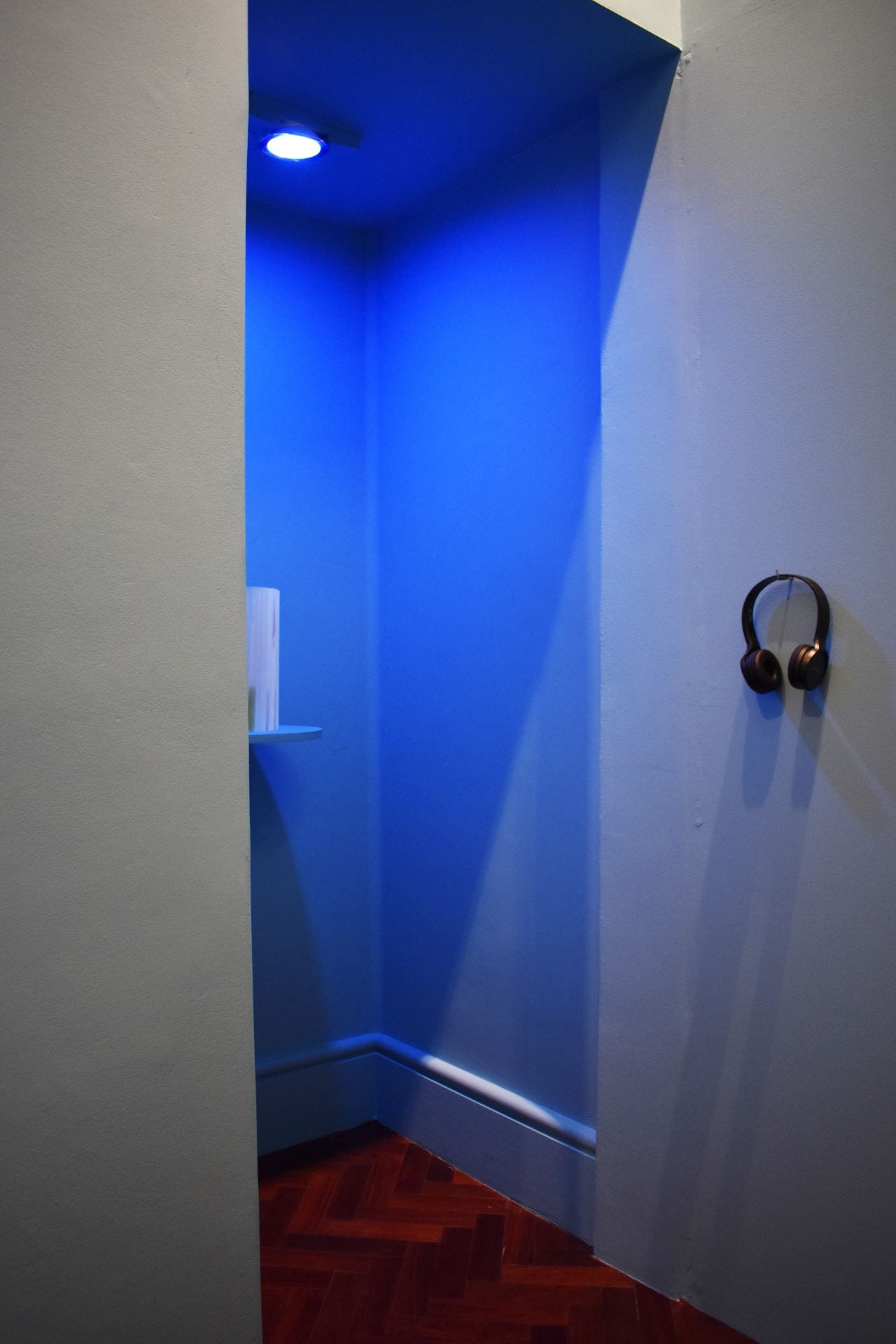 A tall booth space painted in light blue, with blue light shining down inside. A pair of headphones can be seen hanging on a hook on the right-hand wall.