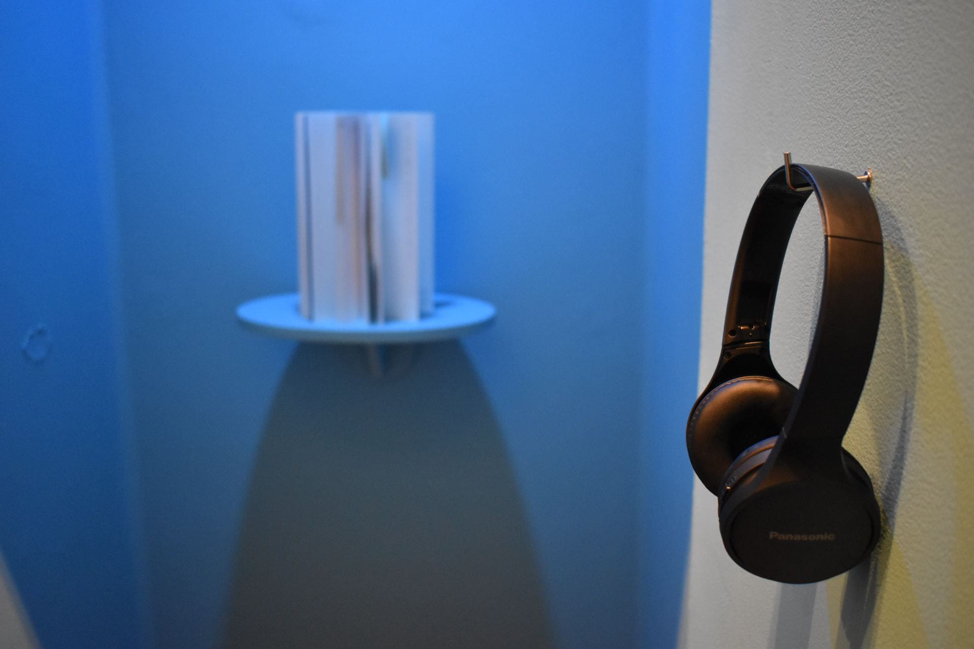 In the foreground, a spiral-bound book is arranged in a circular fashion on a round shelf. In the foreground, a pair of wireless headphones rest on a hook on a blue wall.