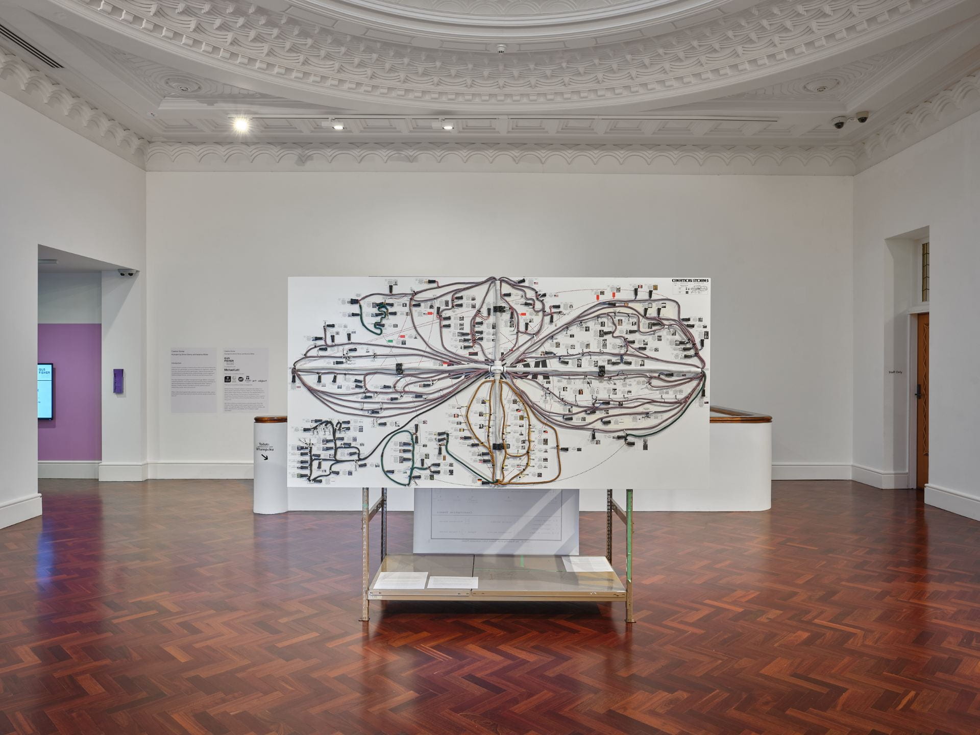 A large board covered in cables and diagrams is attached to a brown metal shelving unit and stands in the middle of a white gallery space.