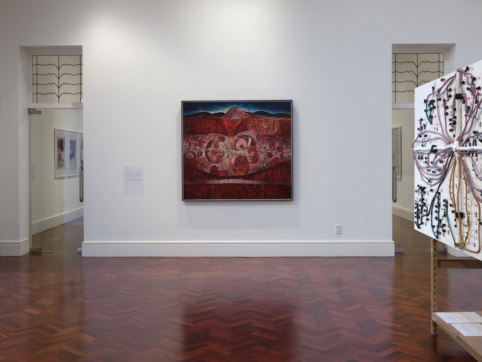 A square painting is hung on a white wall in a large gallery space. The painting depicts a landscape filled with mountains, showing the depths of the earth below. The earth is filled with stylised patterns drawn from Maori kowhaiwhai.