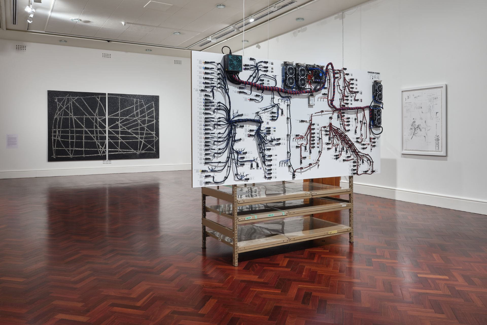 A large board covered in cables and diagrams is attached to a brown metal shelving unit and stands in the middle of a white gallery space.