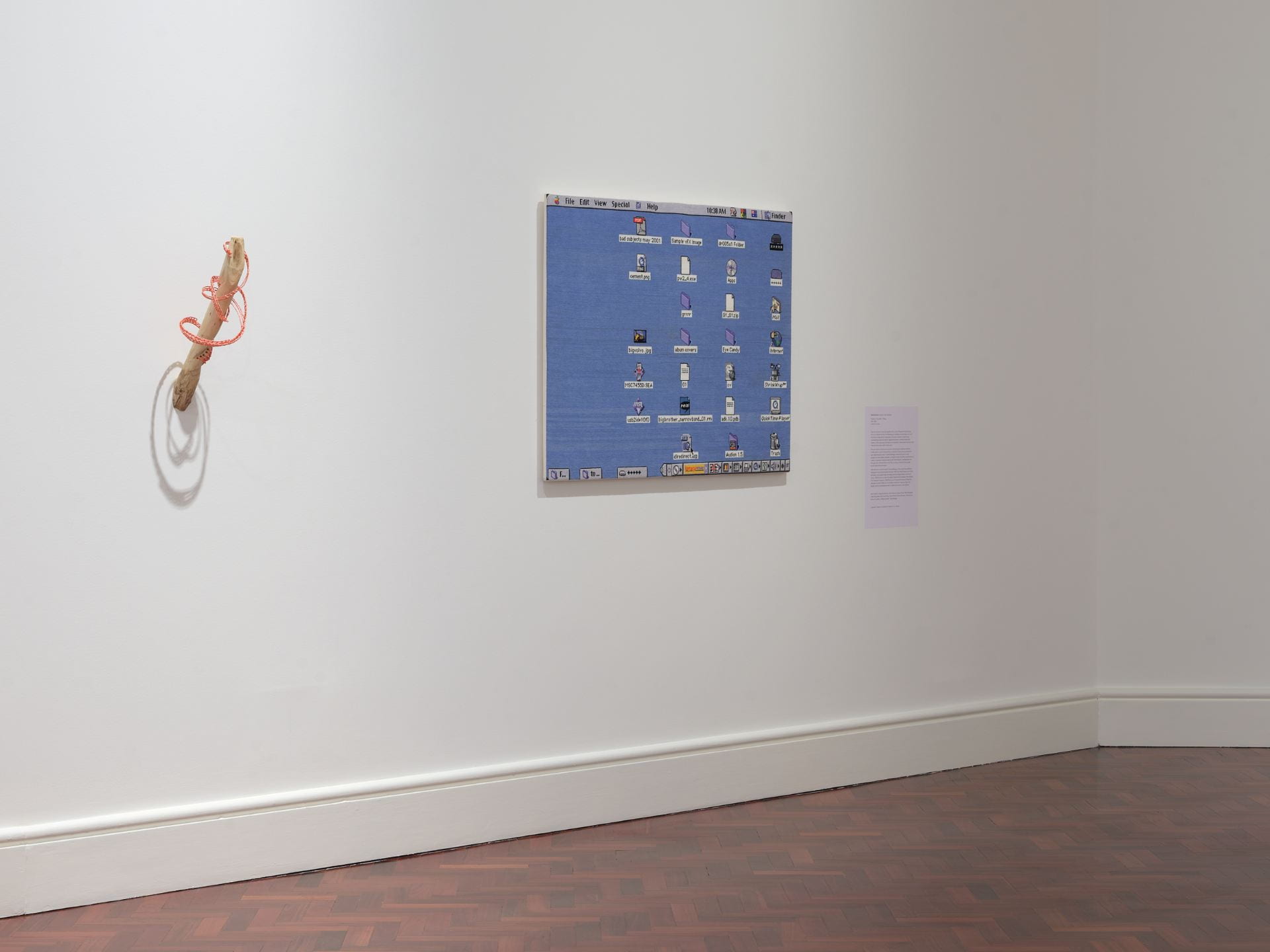 A sculpture of a stick with woven fabric encircling is mounted at an angle onto a white gallery wall. To its right hangs a large embroidery of an old 2001 Mac desktop.