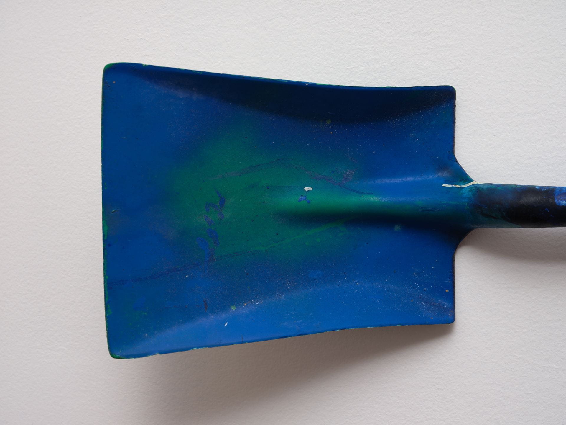 Close-up detail of the head of a green plastic spade, covered in bright blue pigment and hung on a white gallery wall.