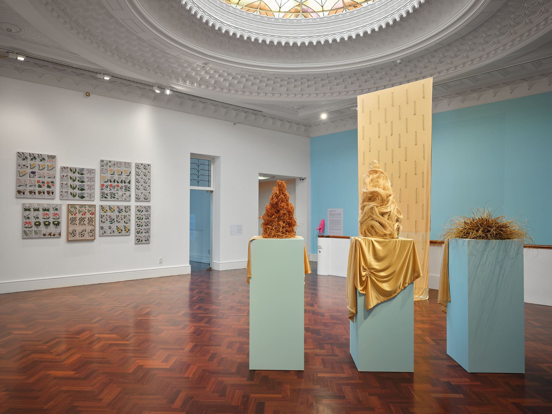 Three tall wigs made of plant fibres are arranged on three pale blue plinths, with golden fabric draped over each. In the background are a series of flower paintings on the left, and a long golden cloak hanging from the ceiling against a pale blue wall on the right.