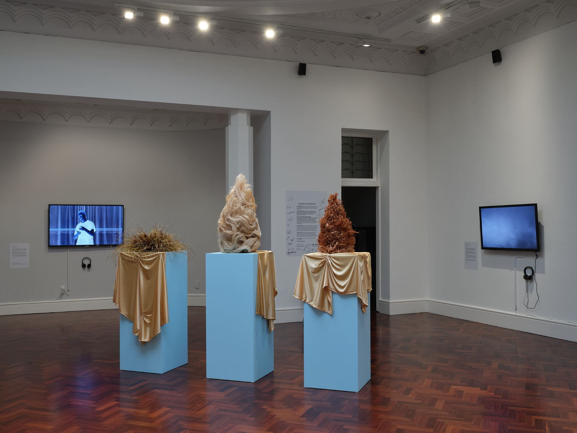 Three tall wigs made of plant fibres are arranged on three pale blue plinths, with golden fabric draped over each. In the background, two TVs mounted on adjacent walls play films.
