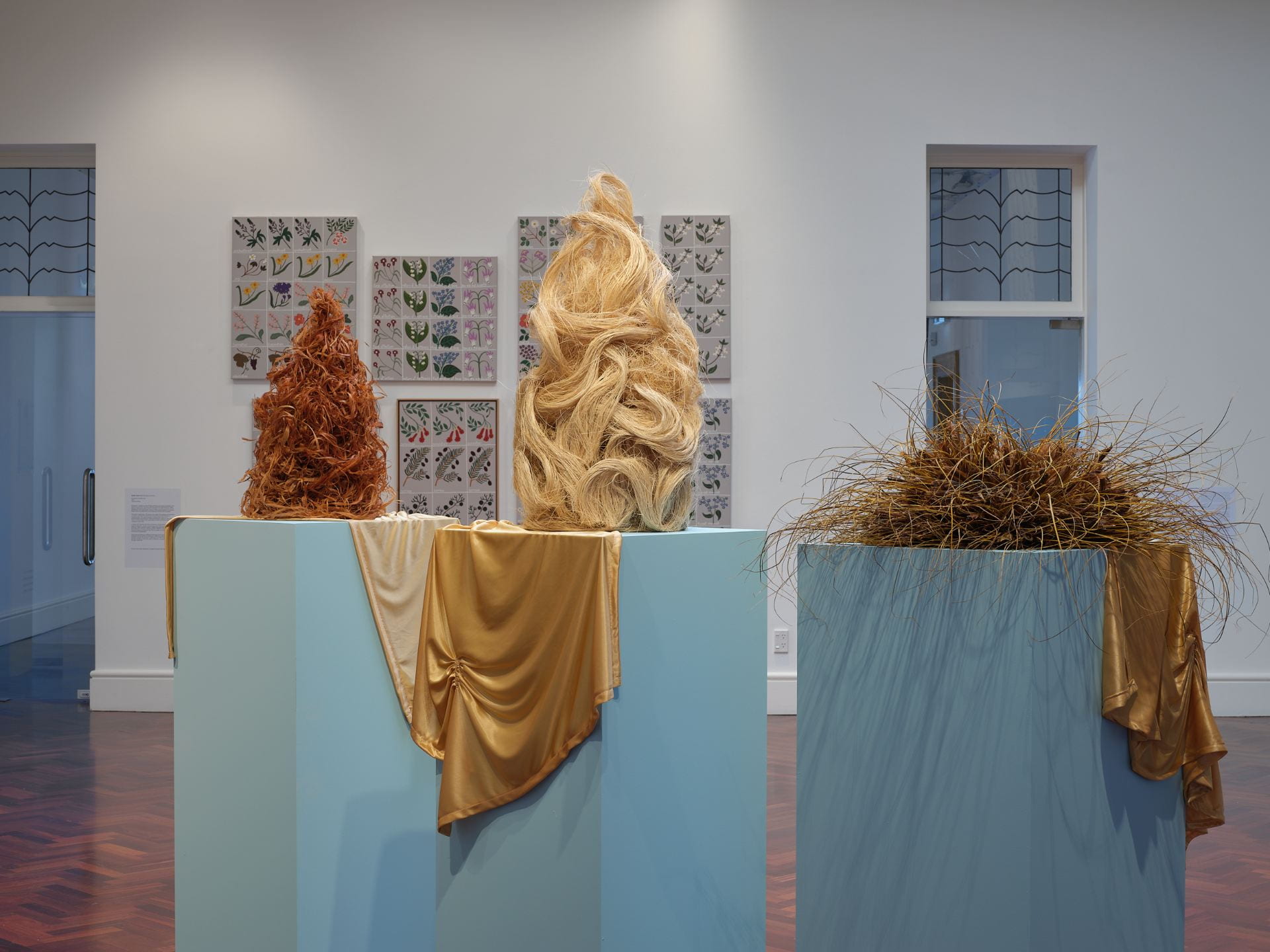 Three tall wigs made of plant fibres are arranged on three pale blue plinths, with golden fabric draped over each. In the background are a series of flower paintings.