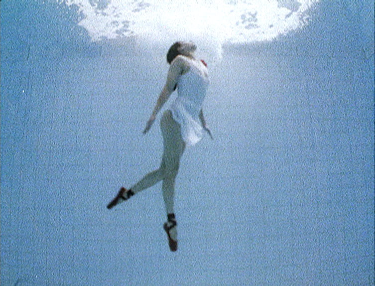 A dancer wearing red pointe shoes is suspended underwater in a huge swimming pool, floating towards the surface. Her face is upturned towards the light.